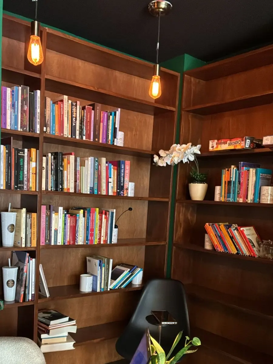 El rincón de lectura del café: libreros de madera llenos de libros hasta el techo, un foco cálido colgando y una silla junto a una mesa pequeña