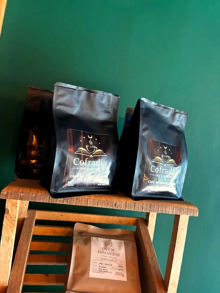 Bolsas de 250 gramos de café Cofradía sobre un banco de madera, frente a la pared verde del local